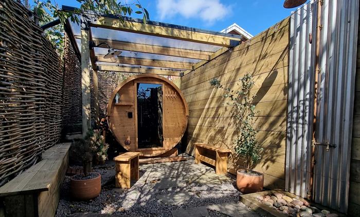 An outdoor, round wooden barrel sauna tucked away under the trees with an outdoor shower and benches for sitting on